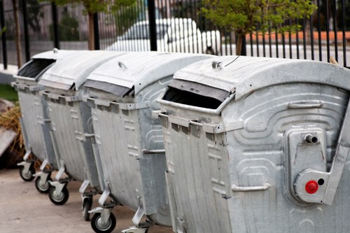 A row of four large, metal wheelie bins positioned on a paved surface, each with a slightly curved lid and metallic finish, showing signs of weathering and minor scratches. The bins are aligned side by side, with their wheels facing outward, and are used for waste or rubbish collection. In the background, there is a metal fence with vertical bars and some trees and parked vehicles beyond, suggesting an urban or residential environment. The scene is evenly lit, with natural light highlighting the metallic texture and subtle shadows cast by the bins. This image reflects typical commercial waste collection equipment, relevant in the context of rubbish removal services in the Wood Green area, as provided by Commercial Waste Removal Wood Green, in a setting that could be part of a business or residential waste management process.