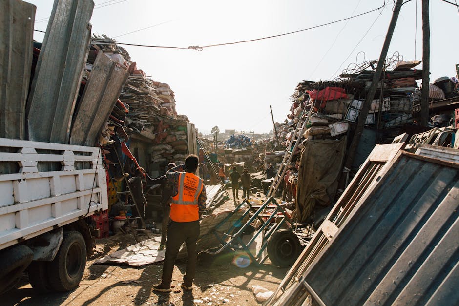 A wide view of a densely cluttered scrapyard or waste collection site with tall, uneven stacks of scrap materials, including rusted metal sheets, discarded wooden planks, and various miscellaneous deb