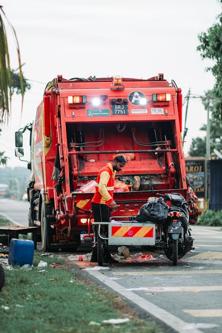 A waste collection worker wearing a high-visibility yellow and orange uniform is operating a large red rubbish truck on a roadside. The worker is leaning forward, using tools to empty or manage waste 